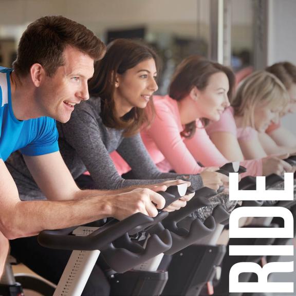 Five adults in line on indoor cycles with wording 'RIDE'